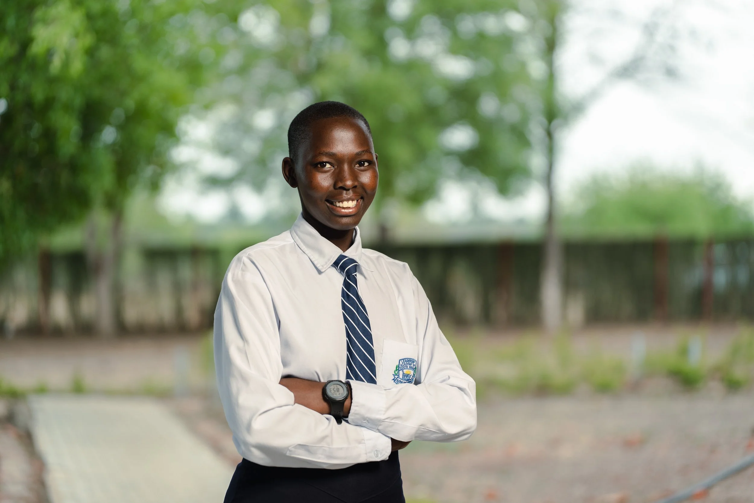A young woman in a school uniform standing outdoors with arms crossed, smiling, with green trees and a fence in the background.