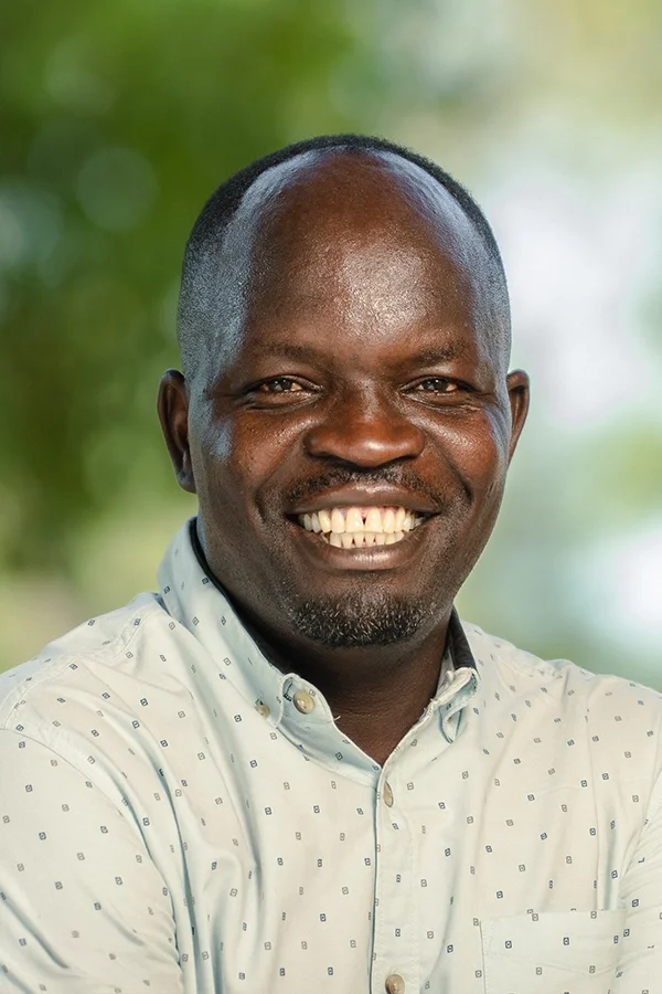 A smiling man with dark skin and short hair, wearing a light-colored button-up shirt, outdoors with blurred green background.