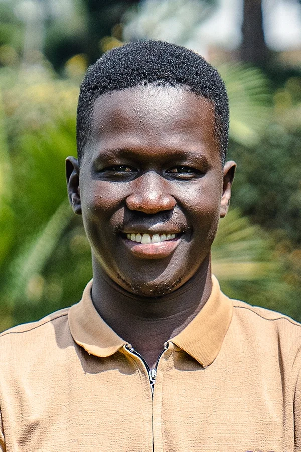 A smiling young man with dark skin, short black hair, wearing a tan zip-up shirt, outdoors with green foliage in the background.