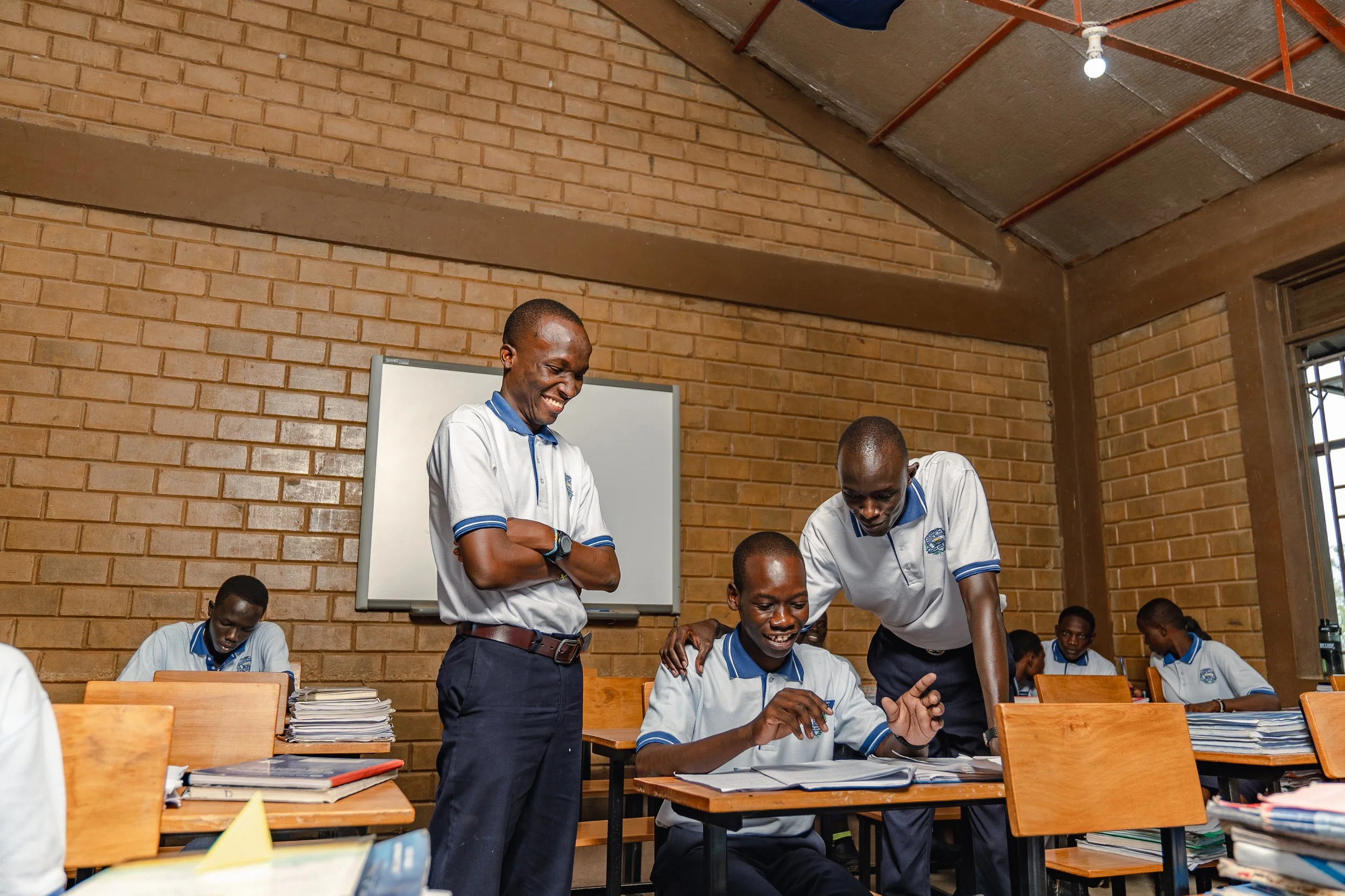 School classroom with male students in uniform, one student shows a book to others, smiling and engaging in learning.