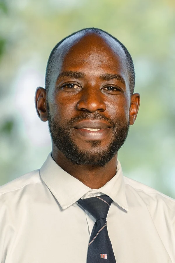 Portrait of a smiling Black man with a beard, wearing a light-colored dress shirt and a dark tie, in front of a blurred outdoor background.