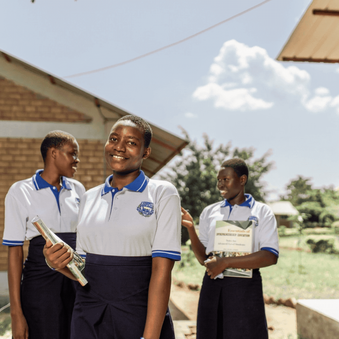 Three girls in school uniforms standing outdoors near a brick building, holding books and smiling.