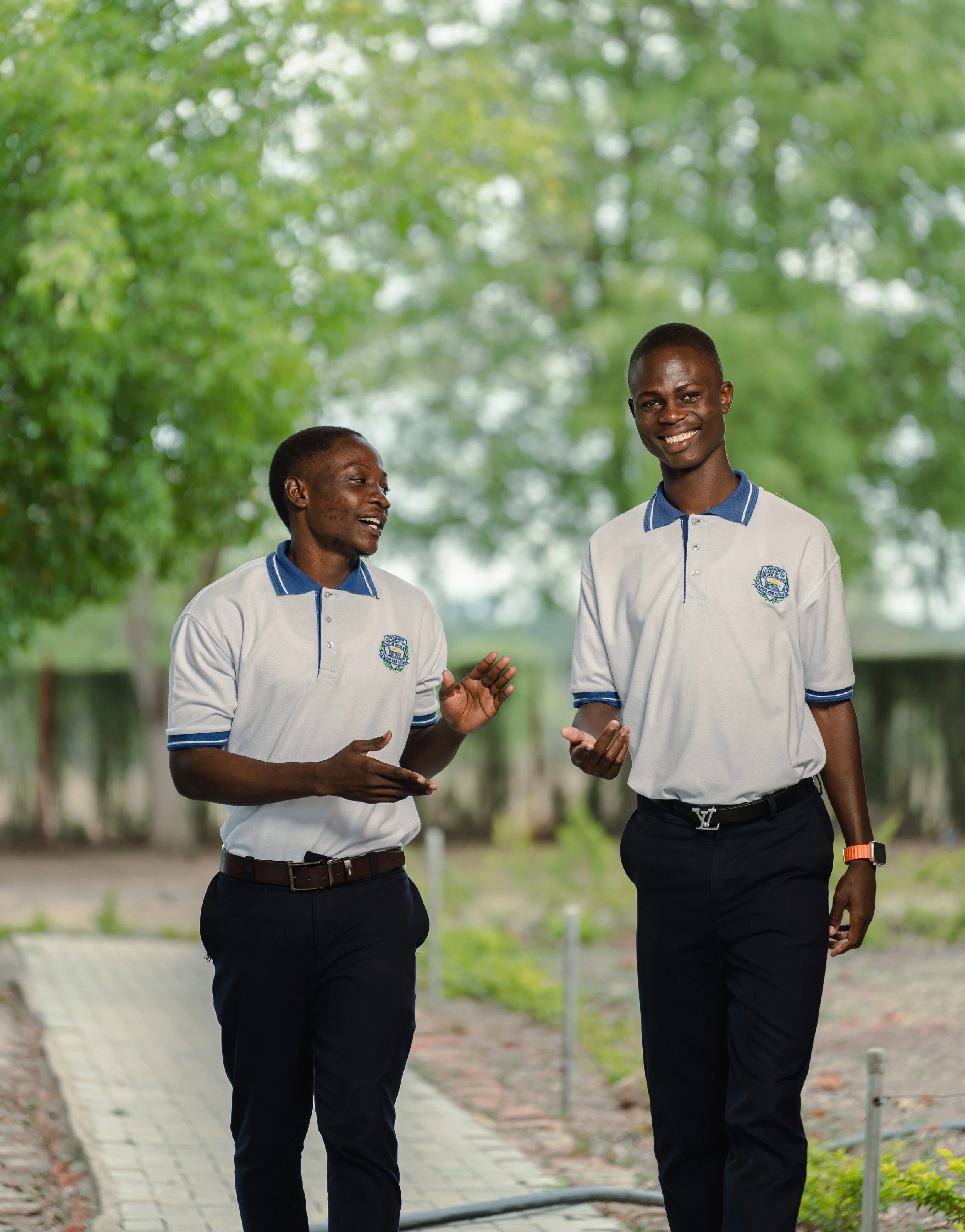 Two men in matching white polo shirts with blue collars and dark pants walking and talking outdoors with green trees in the background.