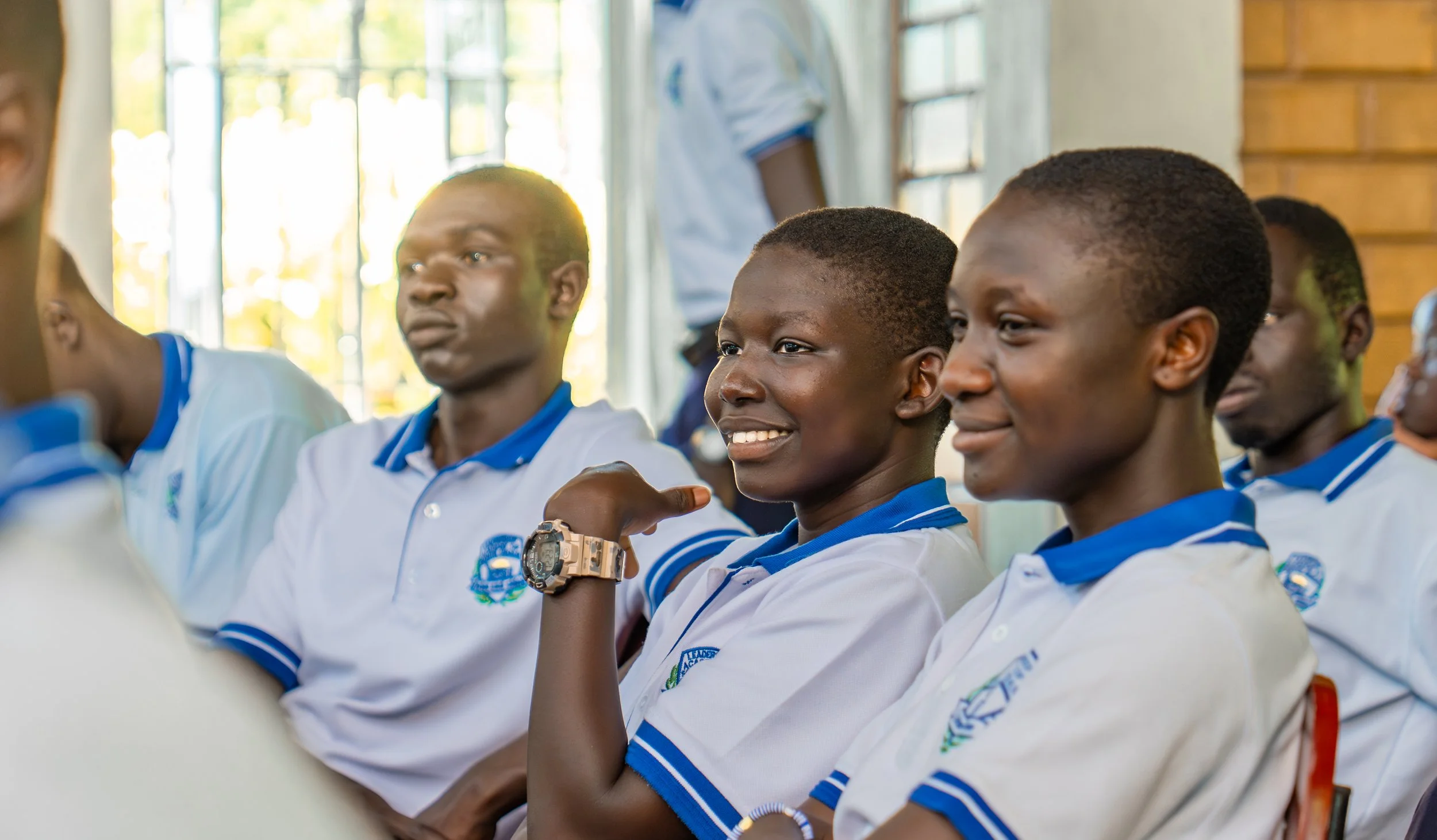 Young students in a classroom, wearing white shirts with blue collars, sitting attentively and smiling. Some students are looking forward, others are looking at something off-camera. The classroom has a large window with sunlight streaming in.