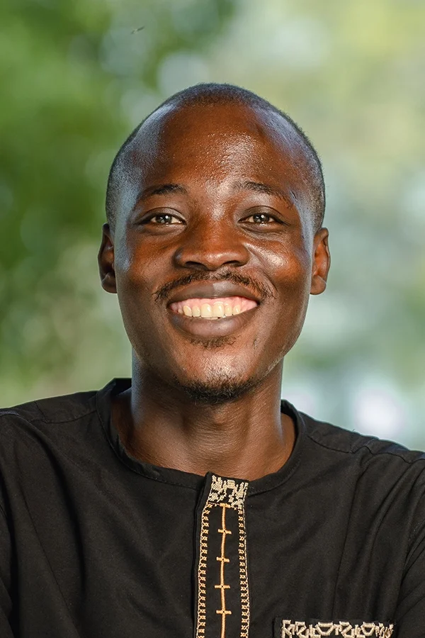 Smiling young man with short hair and a mustache, wearing a black shirt with gold embroidery, outdoors with a blurred green background.