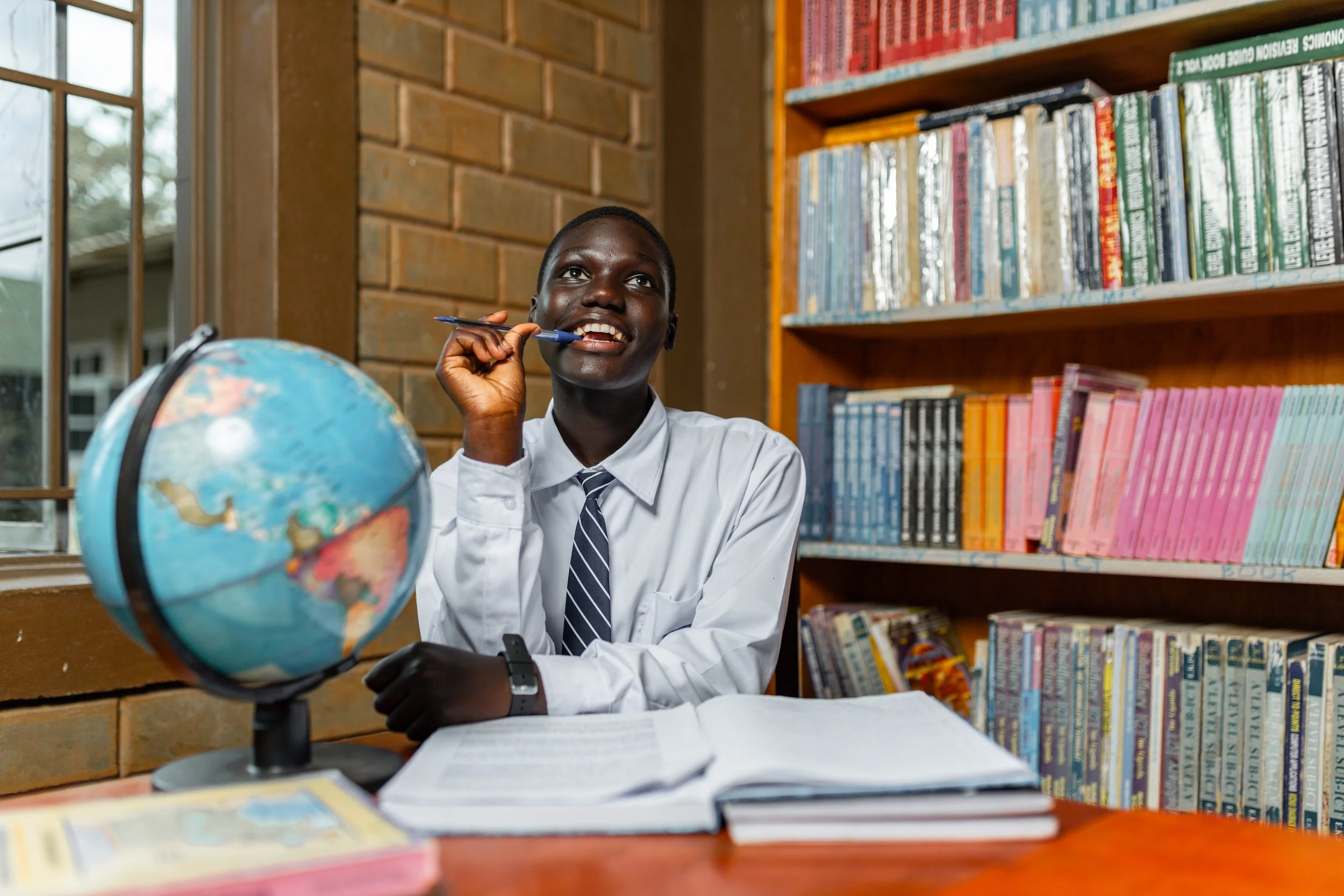A young boy in a school uniform sitting at a wooden desk, with an open book and a globe, holding a pen to his mouth, smiling, in a library with bookshelves filled with colorful books.