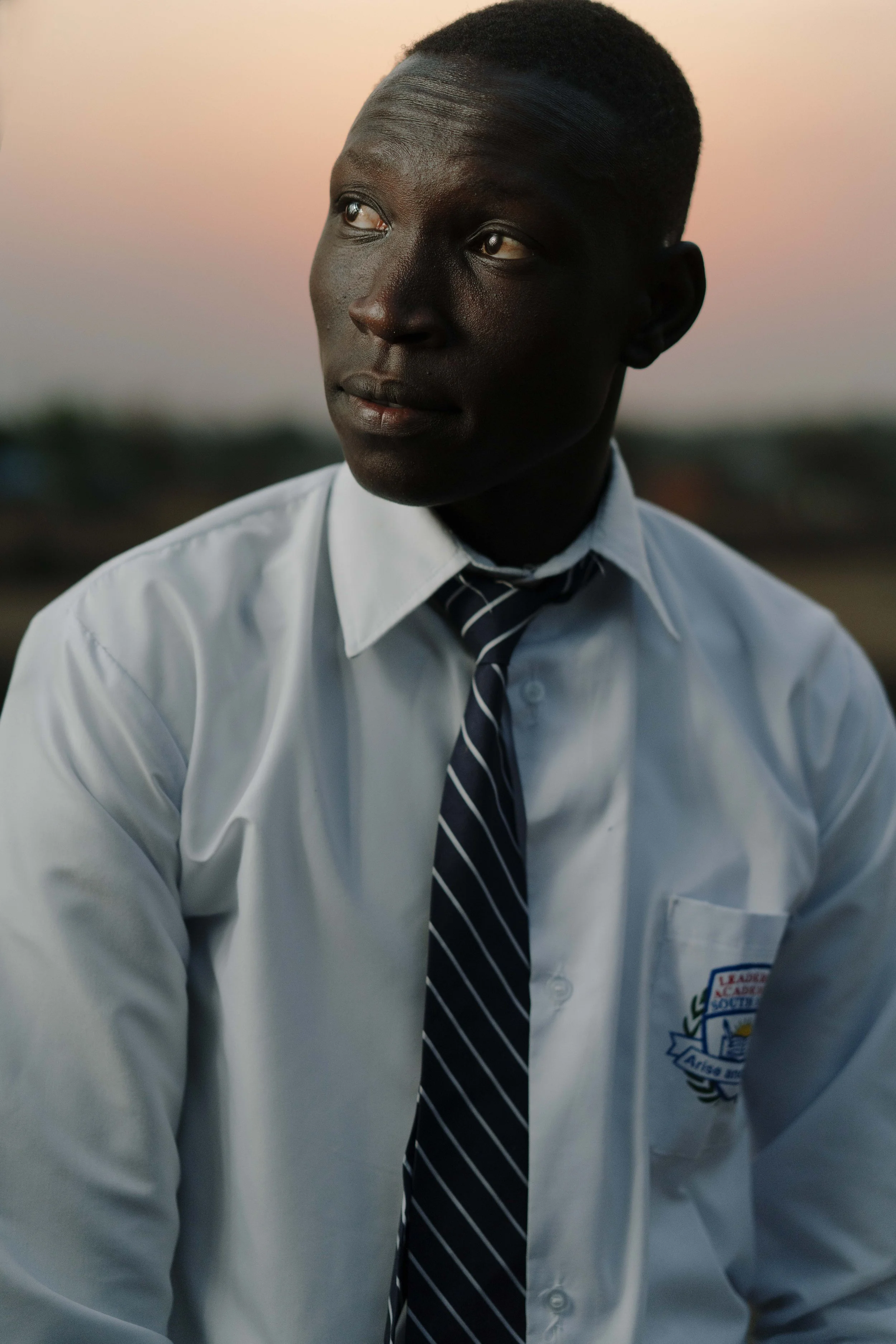 Portrait of a young man wearing a white shirt and a striped tie, looking thoughtfully to the side, with a blurred outdoor background at sunset.