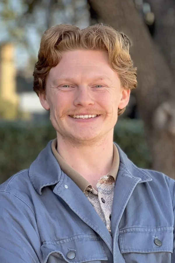 A smiling red-haired man outdoors in front of a tree, wearing a blue jacket and a beige shirt.