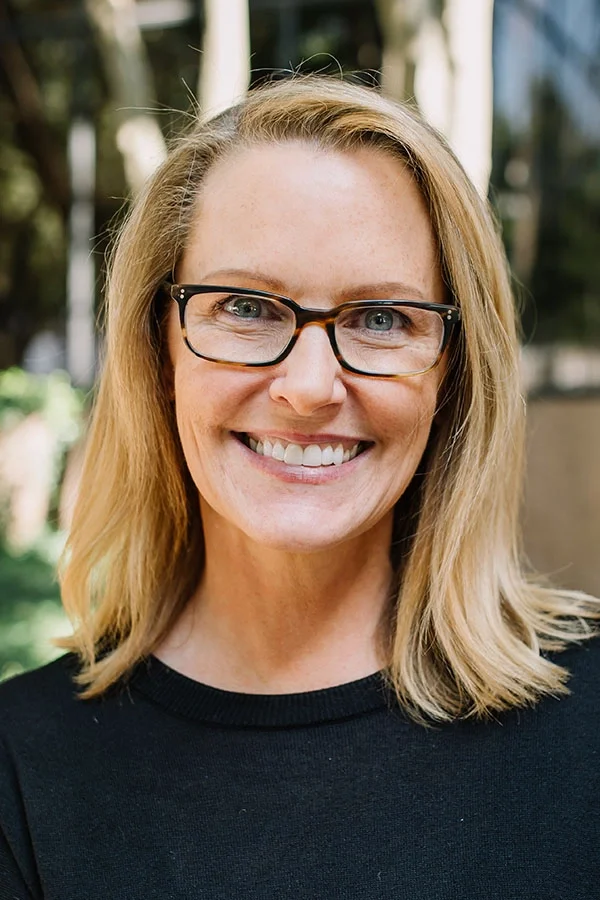 A woman with blonde hair, glasses, and a black shirt smiling outdoors.