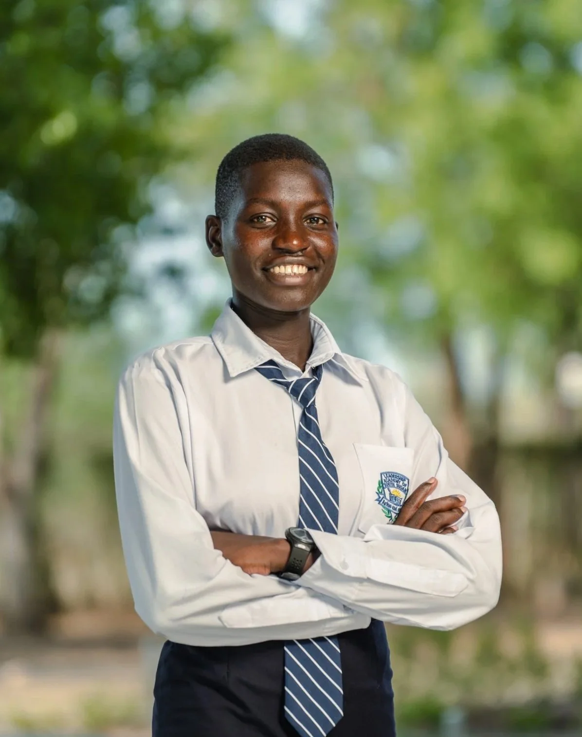 Young girl in school uniform standing outdoors with arms crossed, smiling, with green blurred trees in the background.