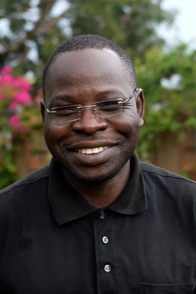 Smiling man wearing glasses and a black collared shirt outdoors with trees and pink flowers in the background.