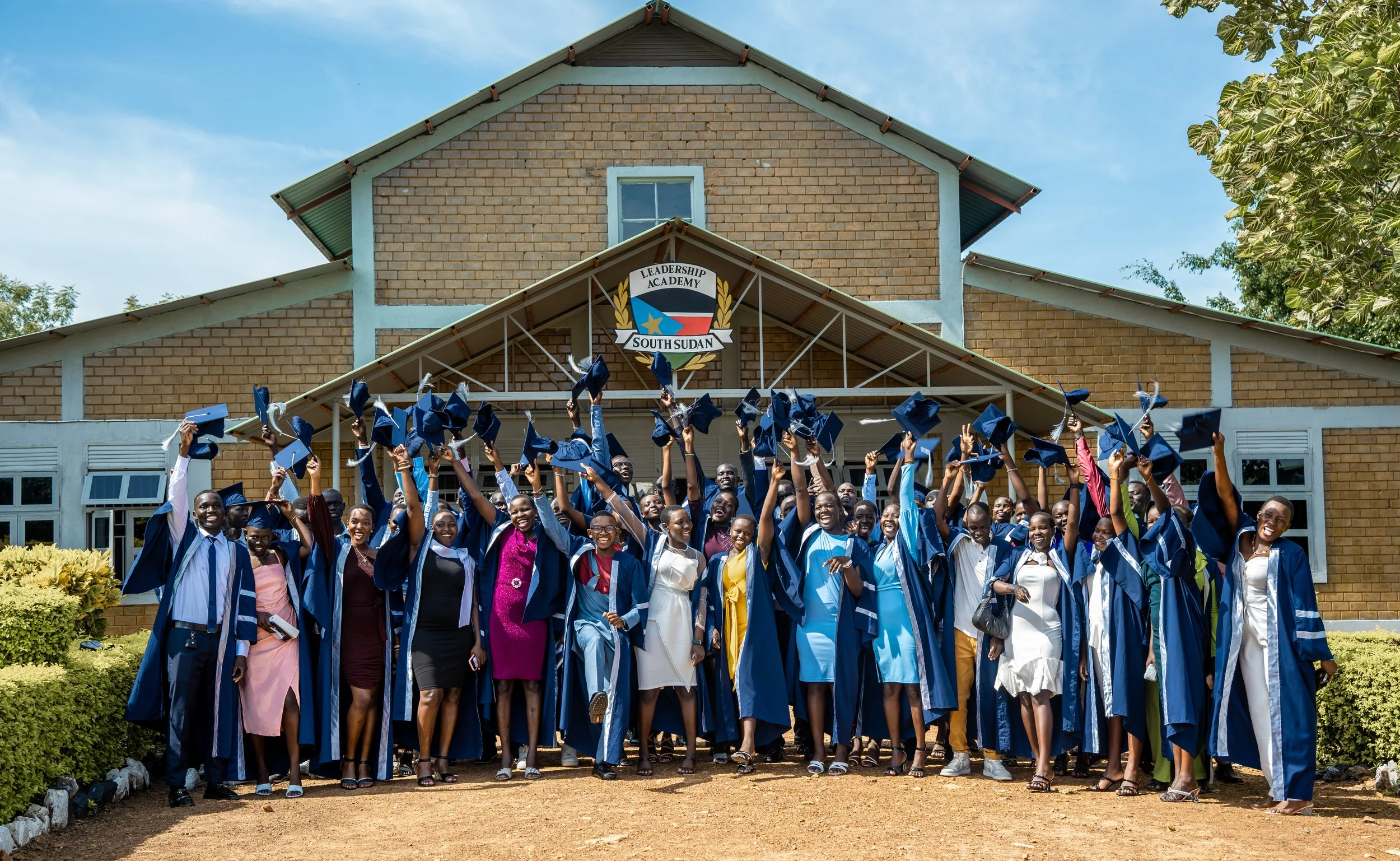 Group of graduates in blue caps and gowns celebrating in front of a building with a sign that reads Leadership Academy South Sudan.