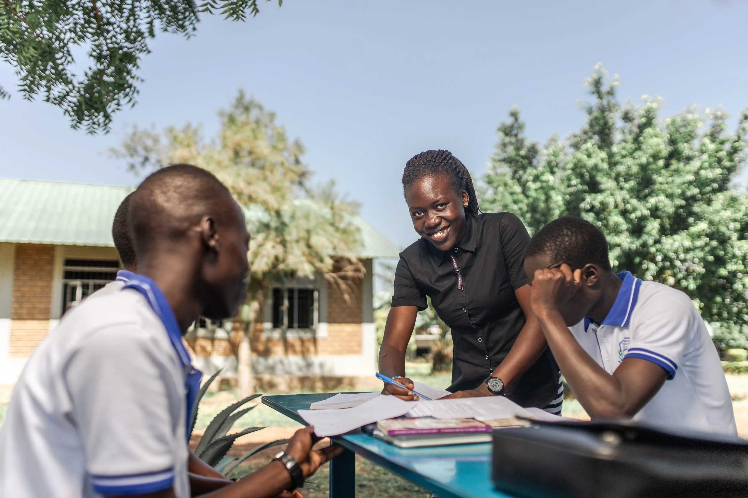 A woman and two young men sitting at a table outdoor, looking at papers and smiling, with trees and a building in the background.