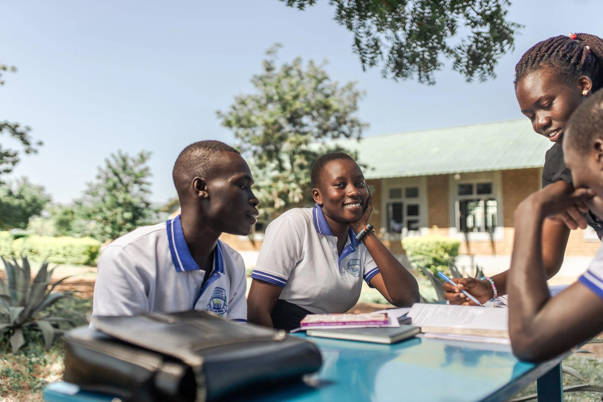 A group of four students sitting at a table outdoors, engaging in a discussion with books and notebooks in front of them, with a school building and trees in the background.