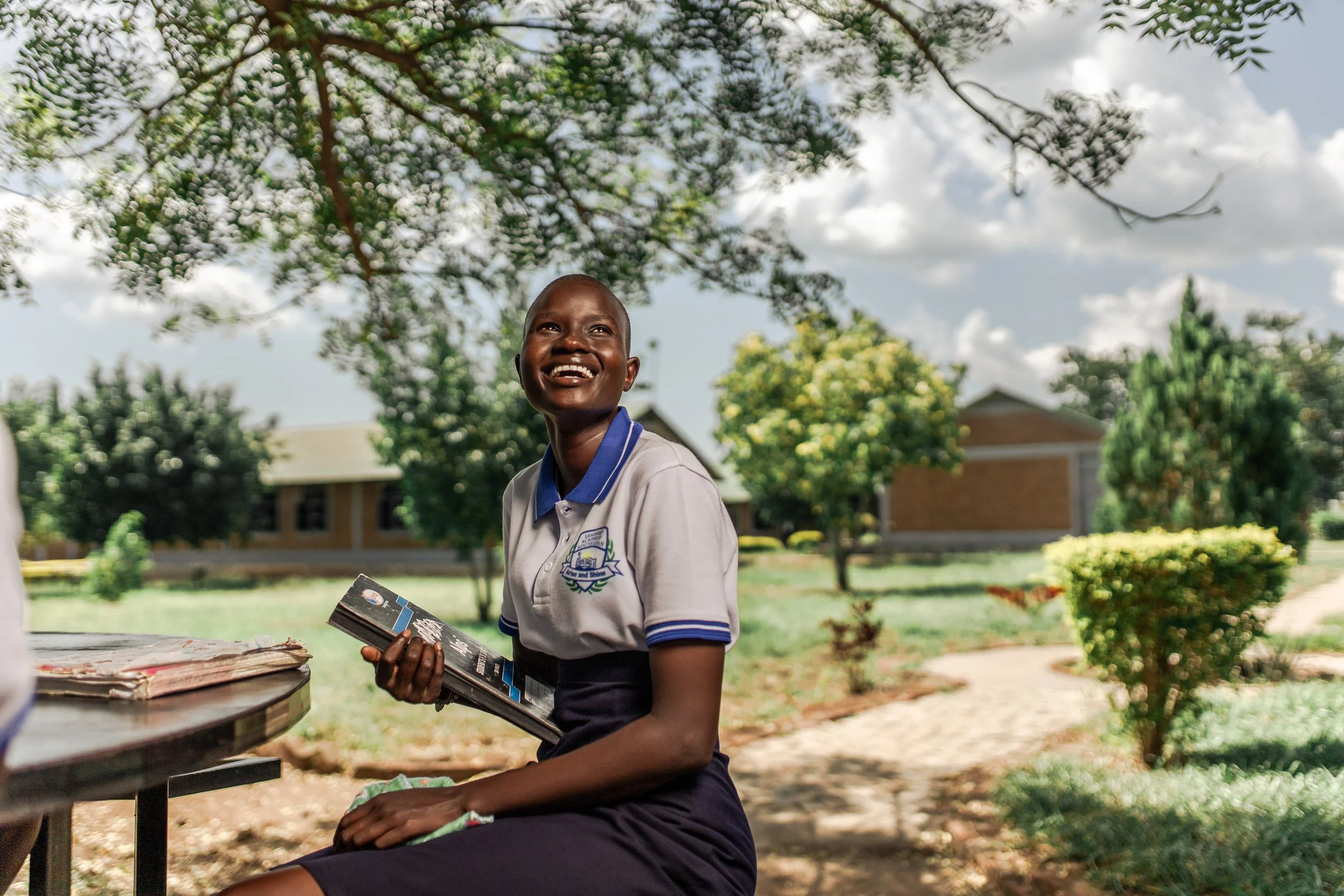 A smiling young girl in school uniform sitting outdoors holding a book, with trees and school buildings in the background under a partly cloudy sky.