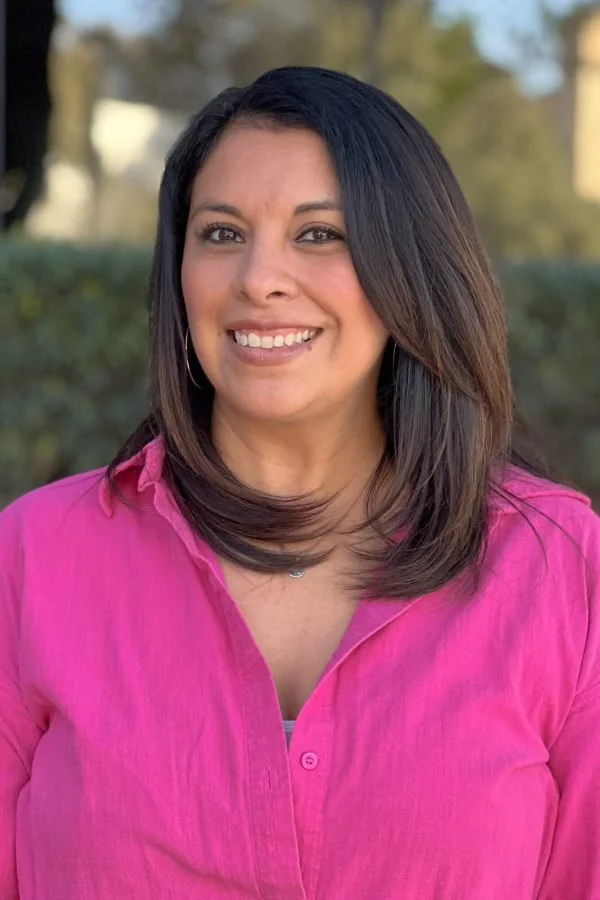 A woman with shoulder-length dark hair, smiling, wearing a pink button-up shirt outdoors.