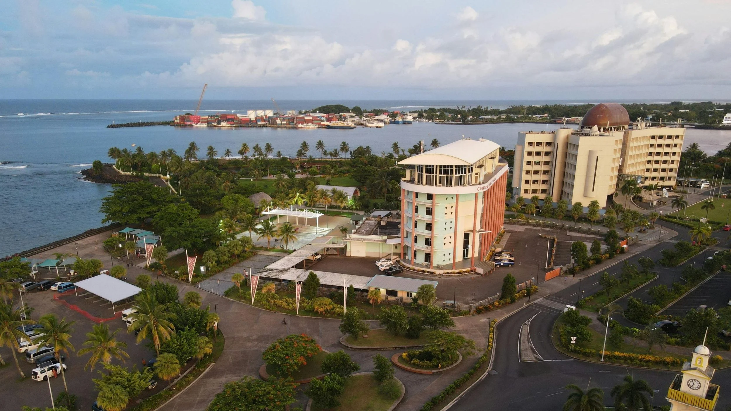 View of a coastal cityscape showing lush greenery, a body of water, and buildings, including a large hotel or office building, with a harbor and shipping containers in the background under a partly cloudy sky.