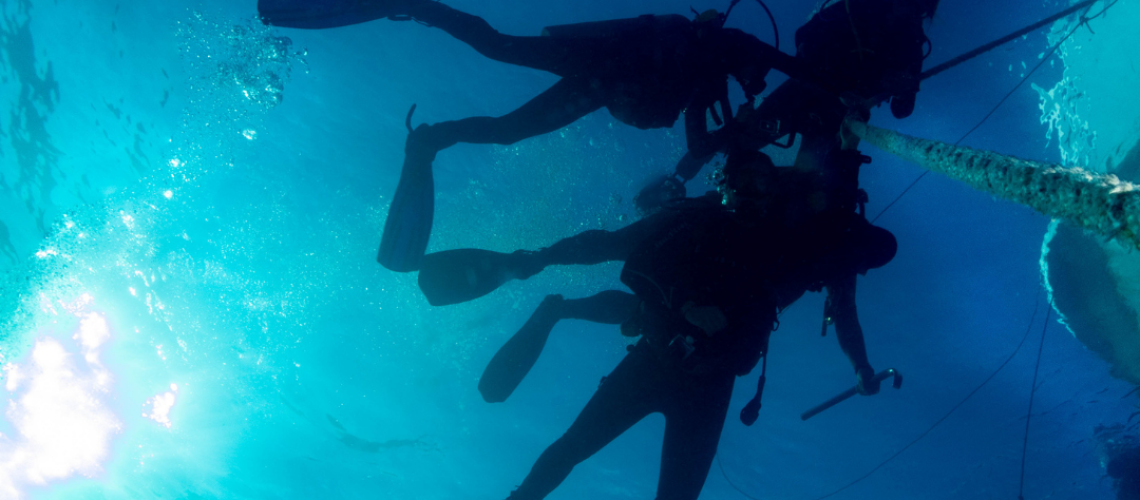 Scuba divers underwater harnessed to a boat, with sunlight filtering through the water.