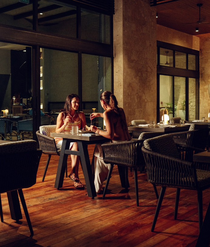 Two women are sitting at a table in a dimly lit restaurant, having a conversation and sharing food, with large windows and a warm wooden floor surrounding them.