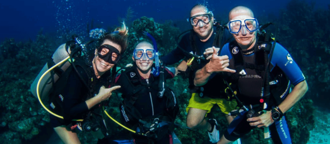 Four scuba divers underwater in a coral reef, smiling and pointing at each other.