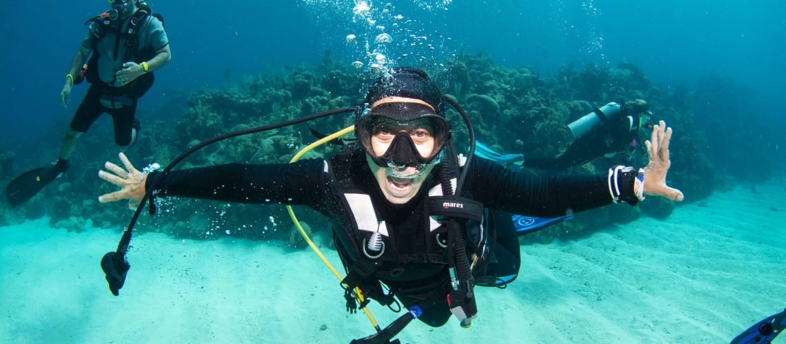 Person scuba diving underwater with outstretched arms, wearing a wetsuit, mask, and snorkel, with another diver in the background.