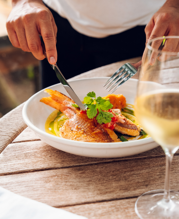 Person using a knife and fork to cut a plated fish dish garnished with cilantro, with a glass of white wine in the foreground.