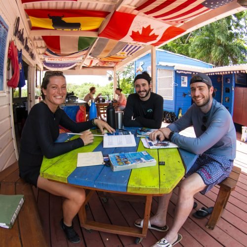 Three people sitting at a colorful outdoor patio table under a roof decorated with international flags, smiling at the camera.