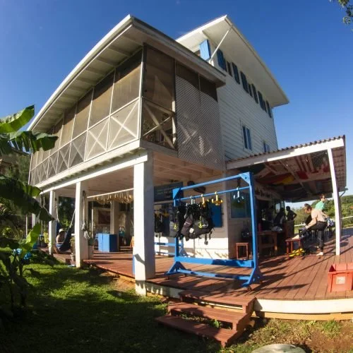 A two-story white house with a wraparound balcony and a porch area with outdoor seating, stores, and a few people at a market or shop.