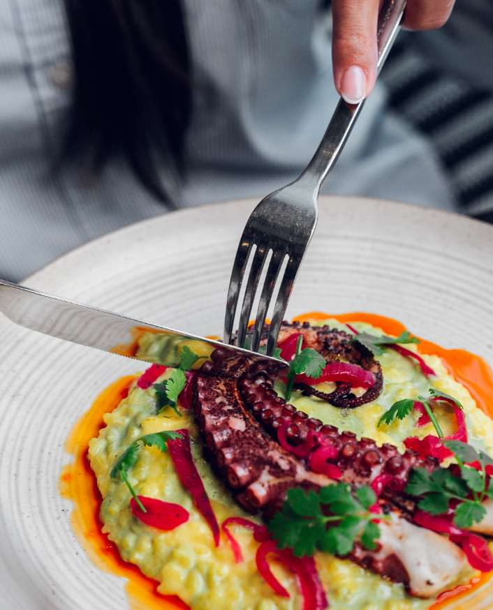 A plate of octopus tentacle on mashed potatoes garnished with red peppers, cilantro, and sauce.