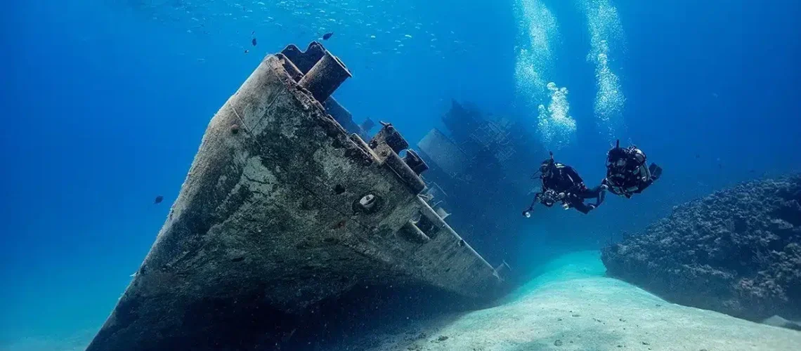 Two scuba divers explore the sunken shipwreck underwater with coral-covered rocks nearby.
