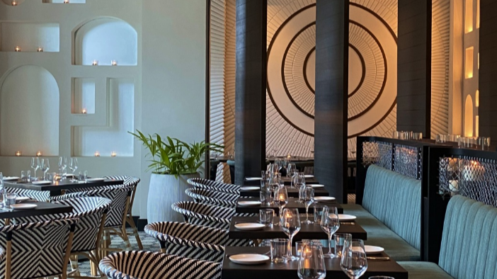 Elegant restaurant dining area with contemporary design. The space features patterned chairs, a long table set with glasses and plates, and ambient lighting. Decorative wall art and plants add to the upscale atmosphere.