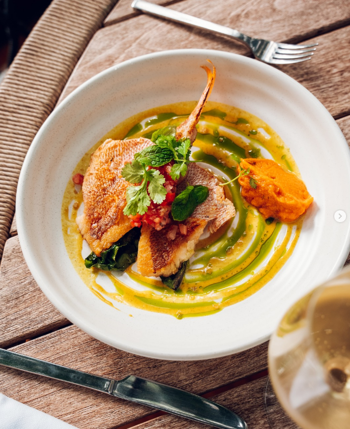 Fried fish fillet garnished with fresh herbs on a white plate, served with a dollop of orange sauce and drizzled with green sauce, on a wooden table with a fork and a glass of white wine.