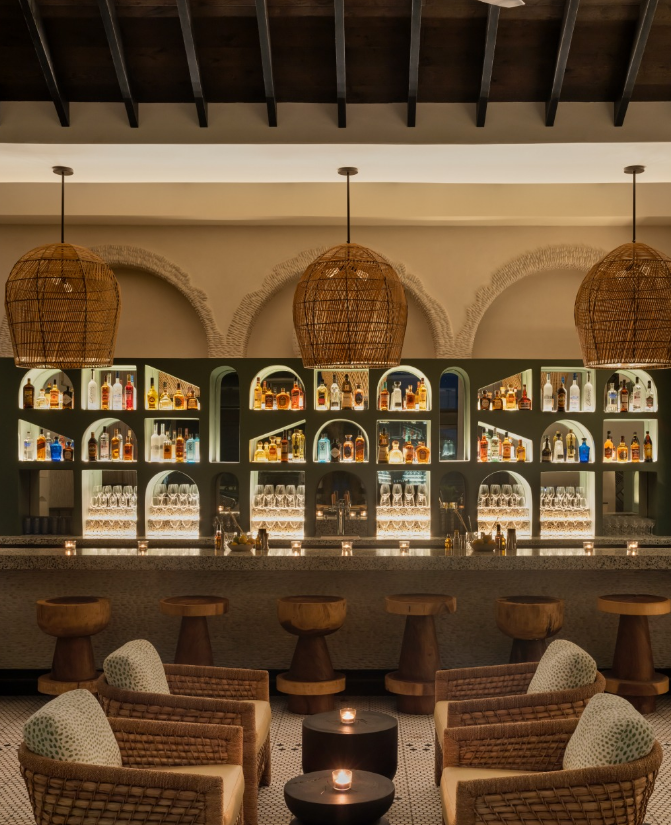 A well-lit bar with illuminated shelves filled with various bottles of alcohol, surrounded by wicker pendant lights and comfortable seating area with armchairs and small tables.