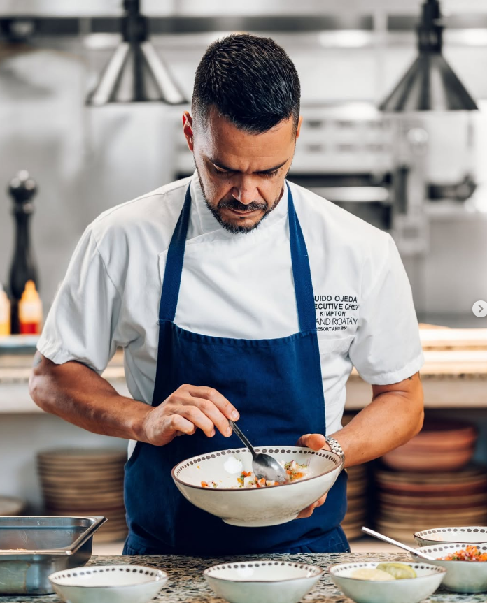 A chef in a white uniform and blue apron preparing food in a professional kitchen.