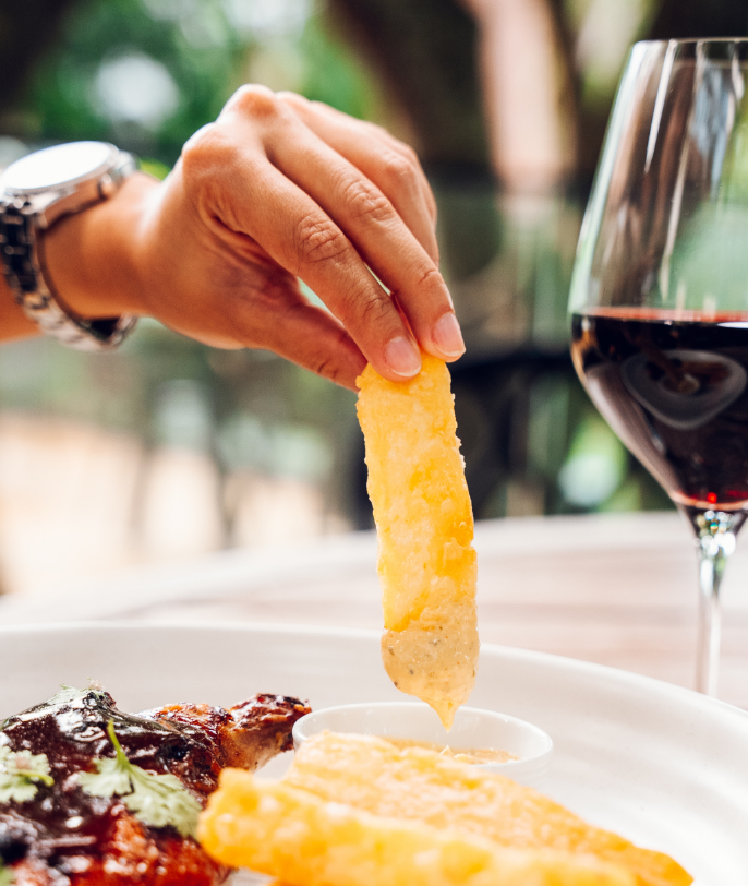Person dipping a fry into a creamy dipping sauce at a table with a glass of red wine and a plate of food.