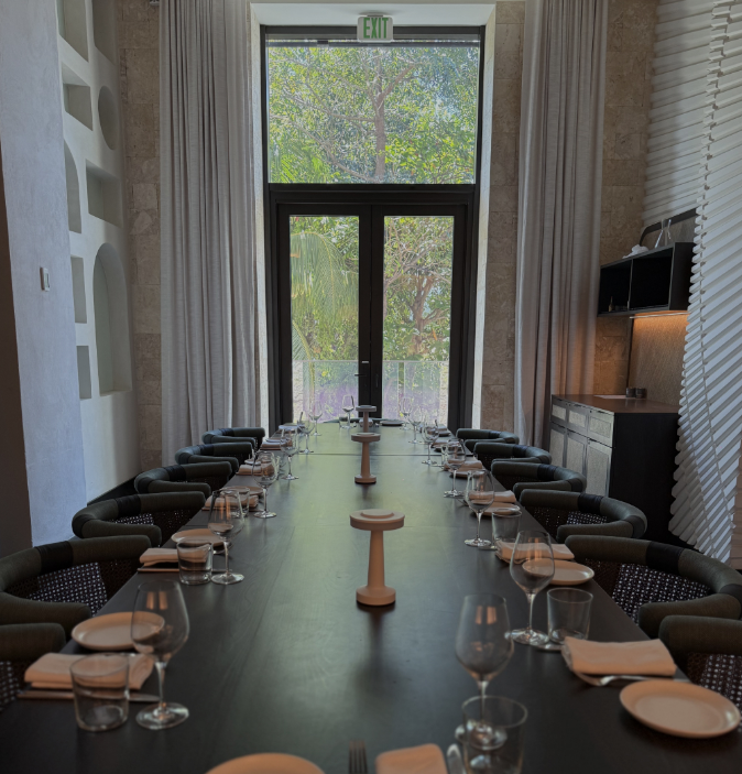 A modern dining room with a long black table set with wine glasses, water glasses, plates, and napkins, surrounded by black chairs, with large windows and curtains, and a view of trees outside.
