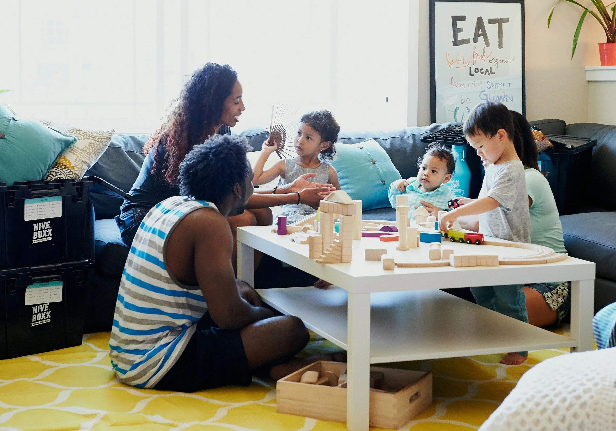 A group of children and a woman sitting on the floor and playing with wooden toy blocks around a white table in a cozy living room.