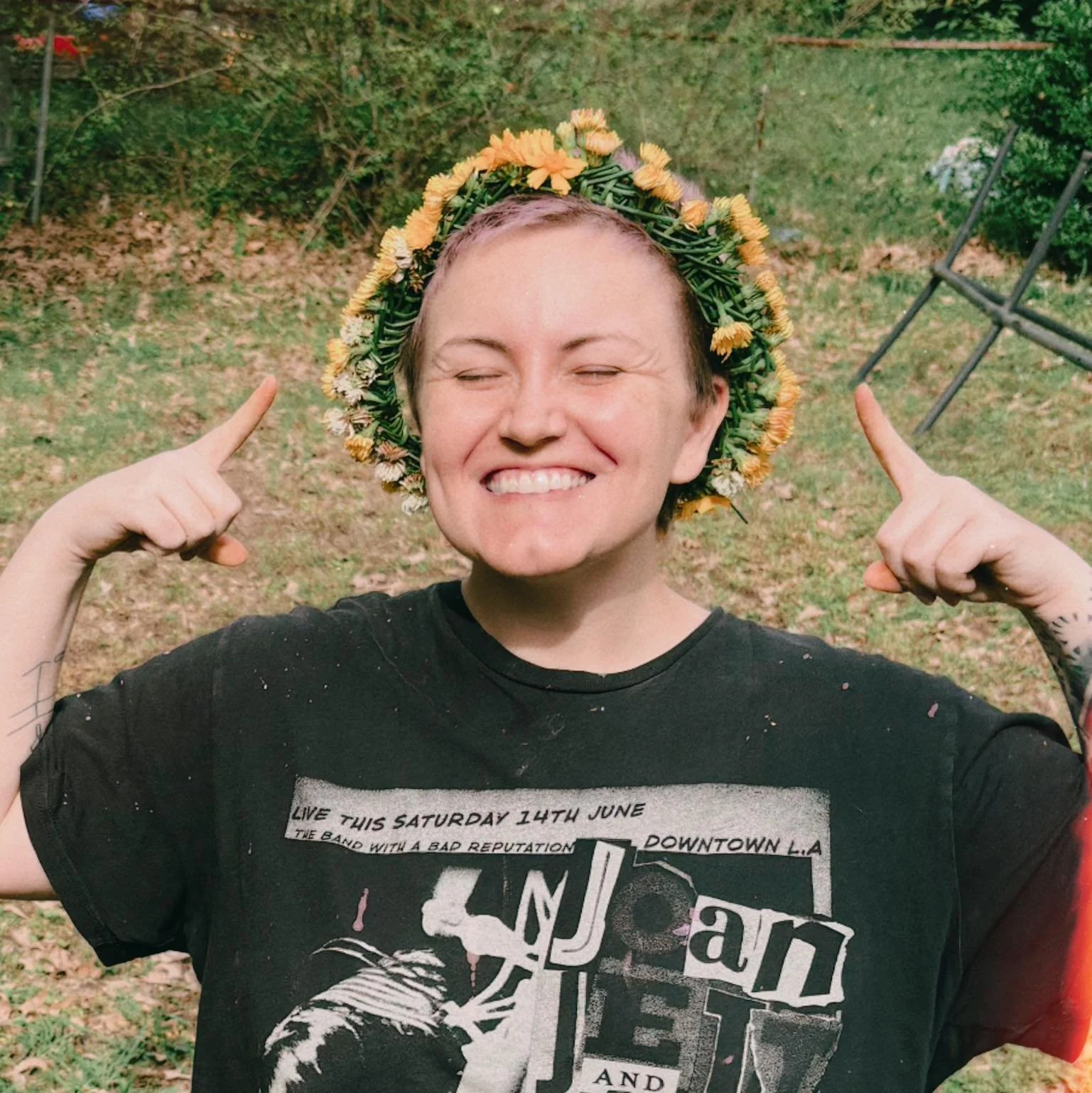 A smiling person with short hair wearing a floral crown and a black T-shirt, pointing to the floral crown while standing outdoors on grass with trees and a metal chair in the background.