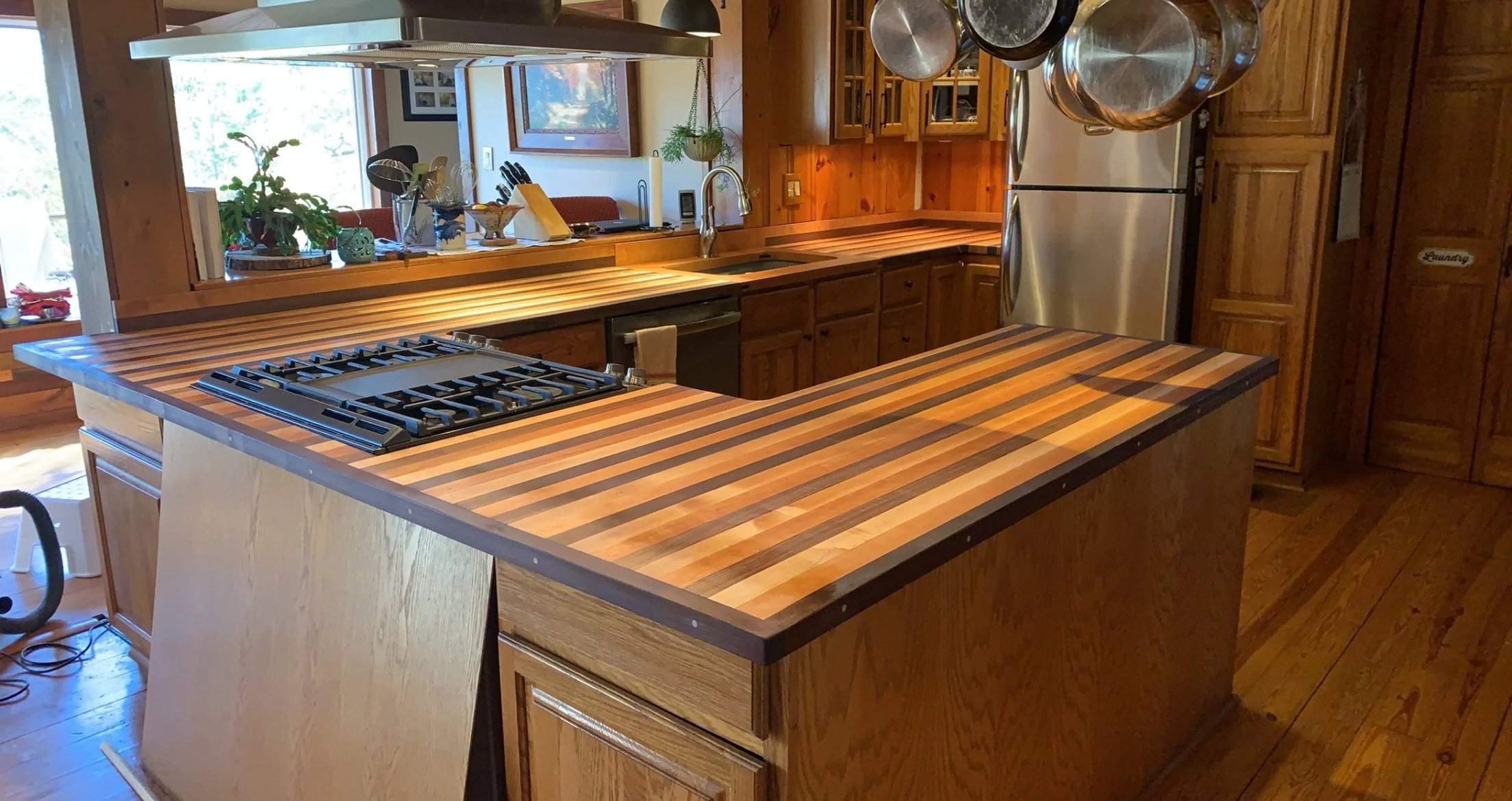 Kitchen with wooden cabinets and a multicolored striped wooden island and countertop. A stainless steel stove is on the island, and a double-door refrigerator is in the background. Pots hang from a rack above the island. A window opens to a dining ar