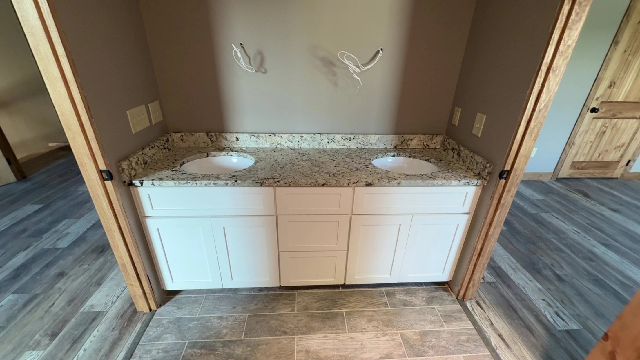 Bathroom vanity with dual sinks, granite countertop, and white cabinets, wall with wiring for lighting, and wood flooring.
