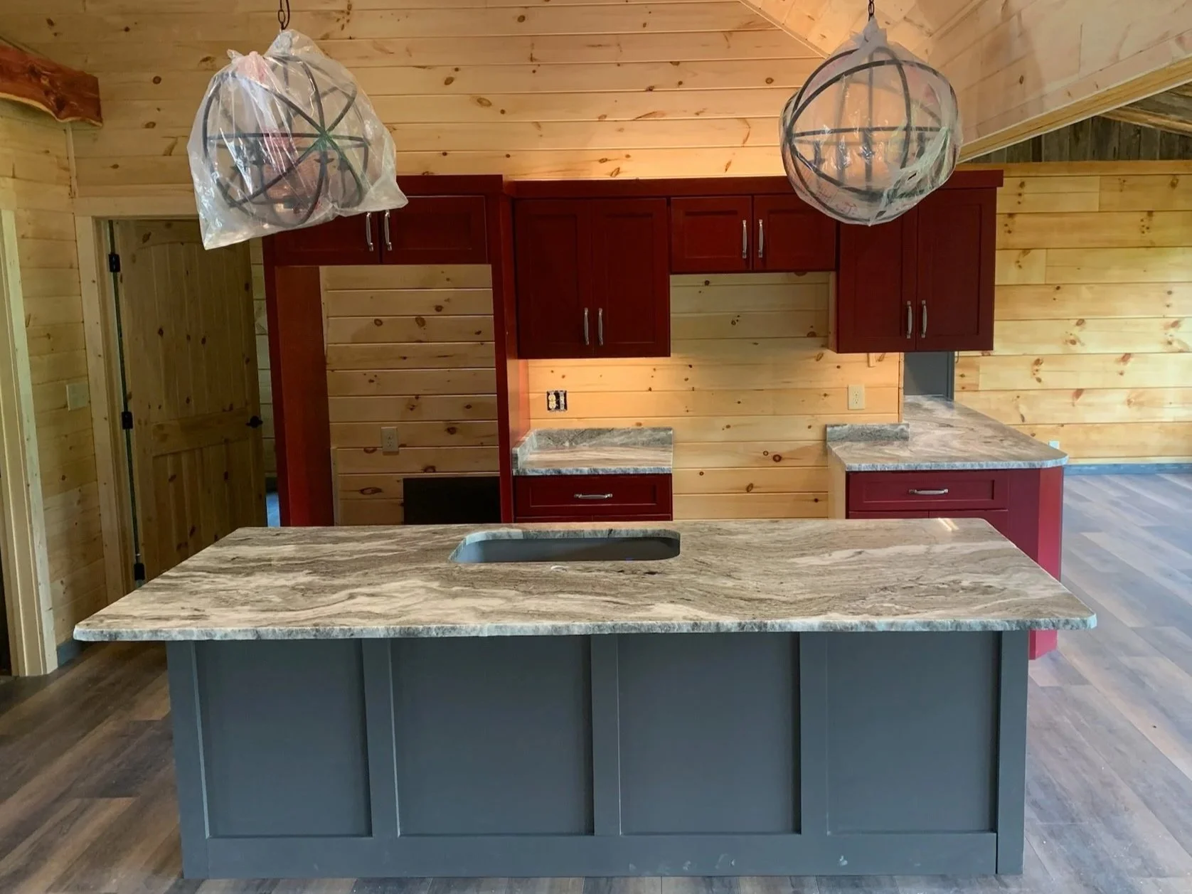 The kitchen features a large island with a gray base and a marbled granite countertop, set in a wood-paneled room with natural light, featuring red cabinets, wood walls, and two decorative globe pendant lights covered in plastic.