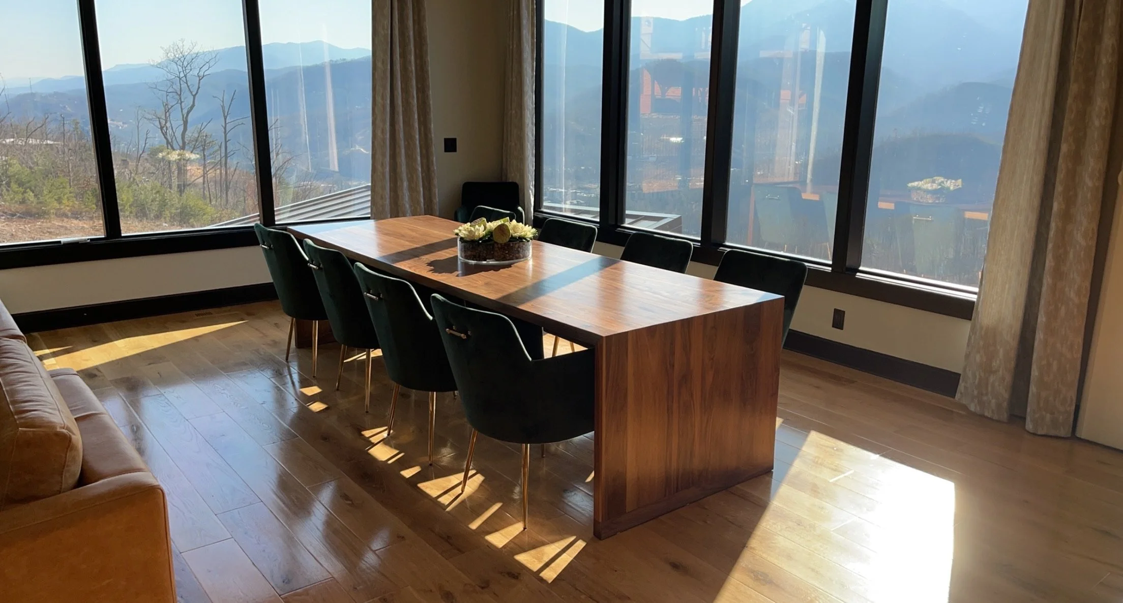 Dining room with a wooden table surrounded by black chairs, sunlight streaming through large windows with a mountain view, and a floral centerpiece on the table.