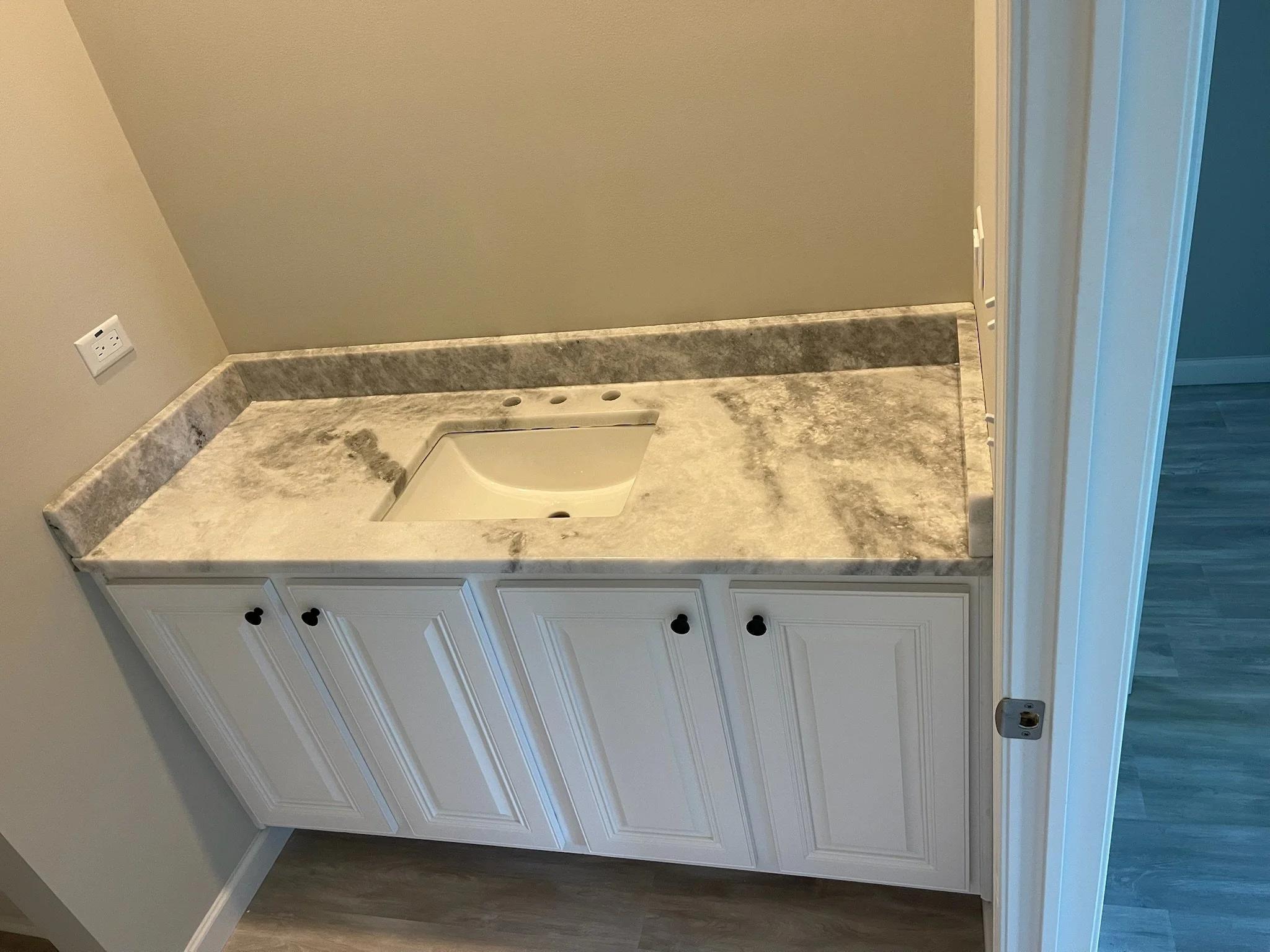 A bathroom vanity with a marble countertop that includes a small undermount sink, white cabinet doors with black heart-shaped knobs, and a wall outlet on the beige wall.
