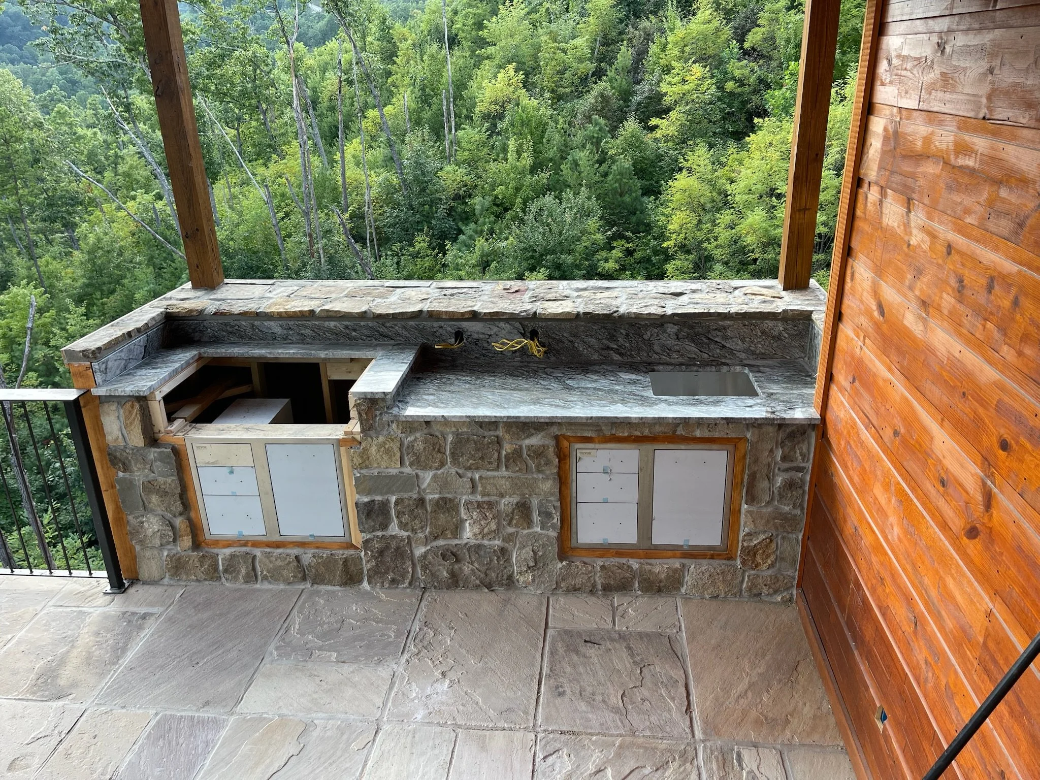 Outdoor kitchen under construction with stone and wood materials, featuring a granite countertop with a sink, an open cabinet space, and windows with plastic coverings, overlooking a lush green forest.