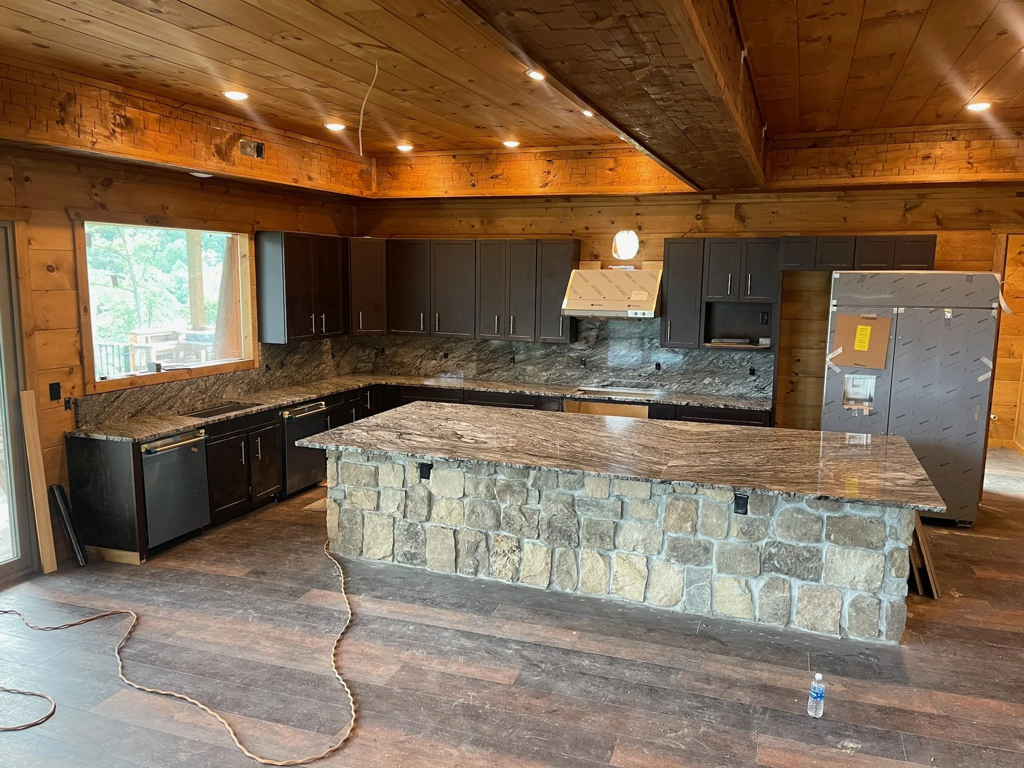 Modern kitchen with wooden walls and ceiling, dark cabinets, marble countertops, a stone island, and stainless steel appliances. A window shows trees outside, and construction tools and materials are present.