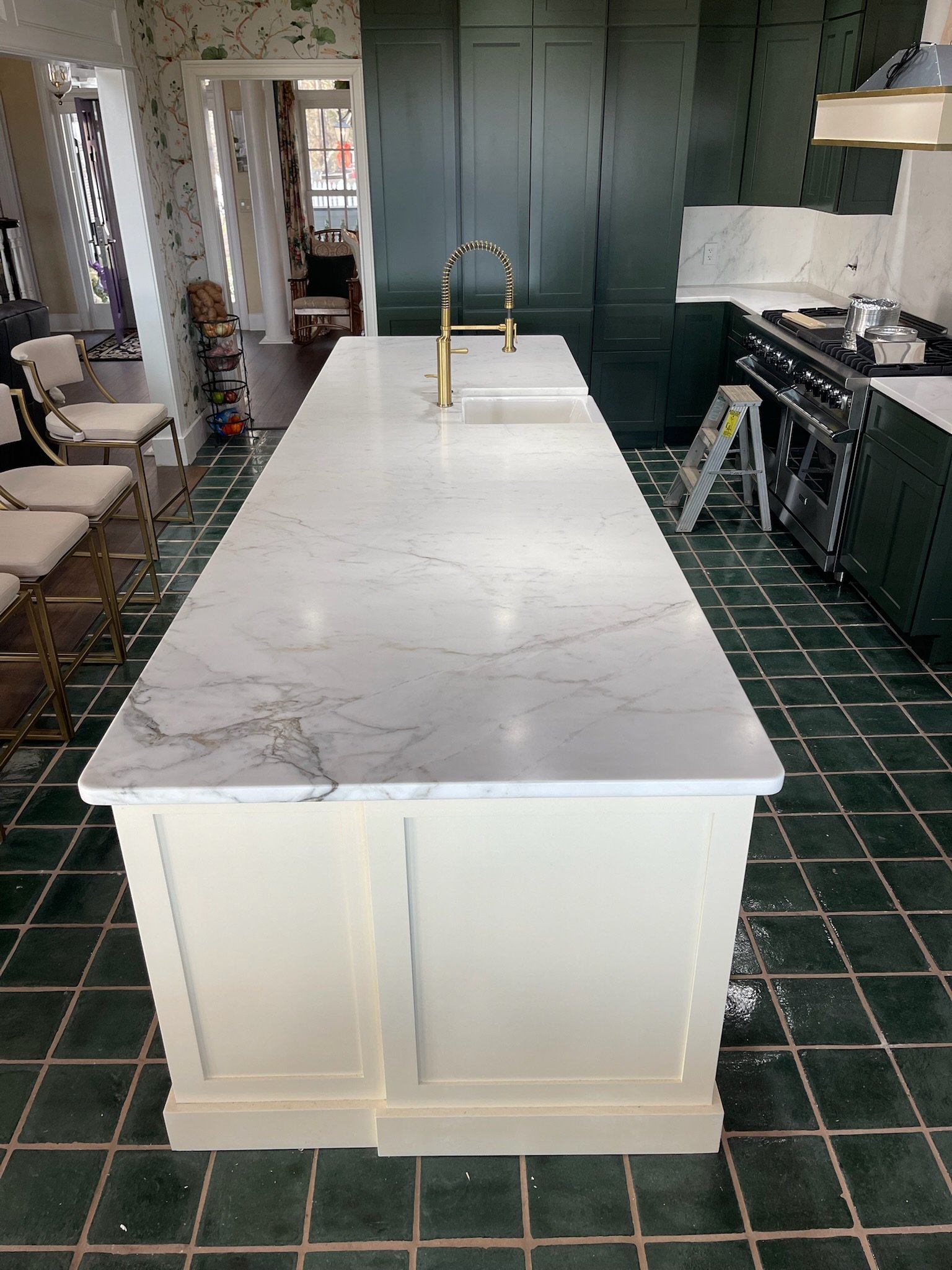 Modern kitchen with white marble island, gold faucet, dark green cabinets, black stove, tiled dark green floor, and view into adjacent rooms.