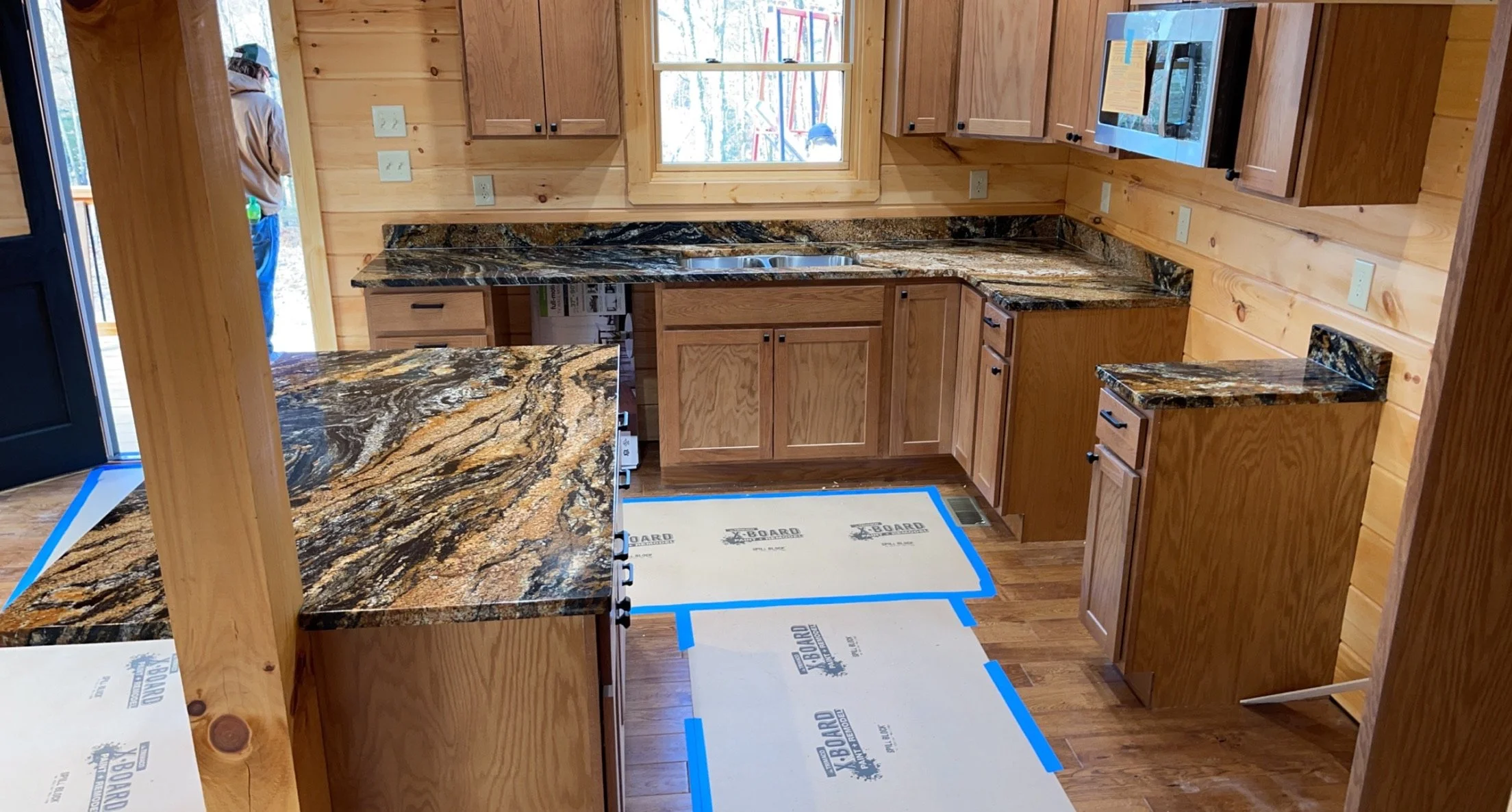 Kitchen with wooden cabinets, granite countertops, a black microwave mounted above the counter, a window behind the sink, and a person standing outside the door to the left.