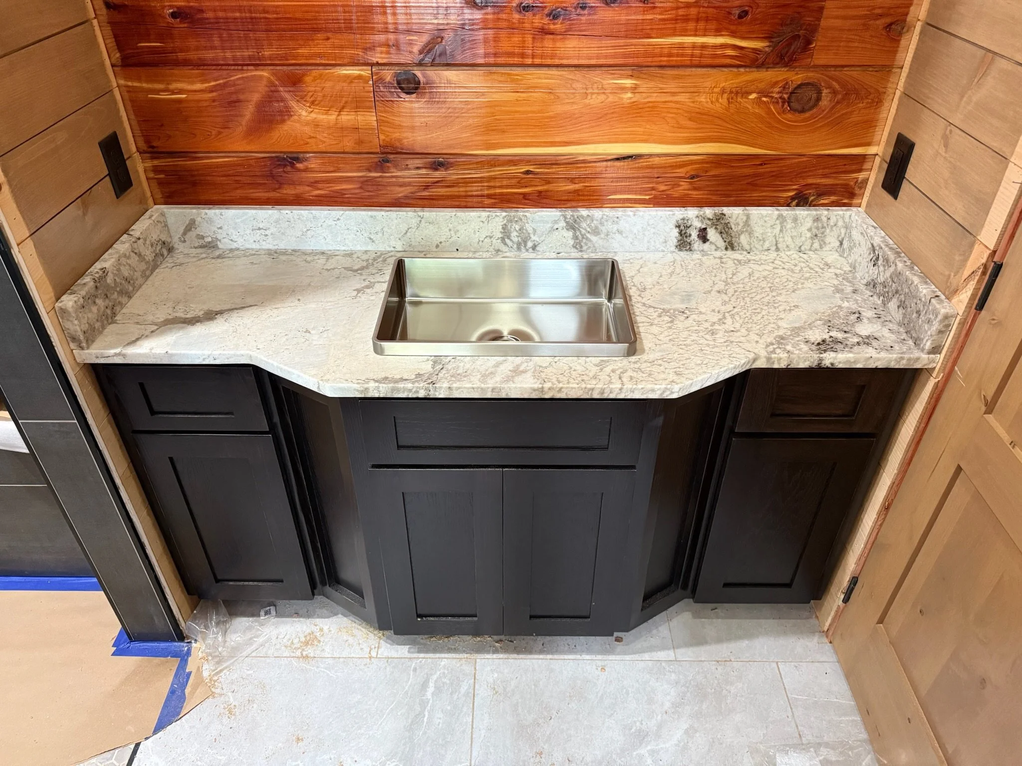 A kitchen counter with a granite surface, black cabinets, a stainless steel sink, and a wooden wall backdrop.