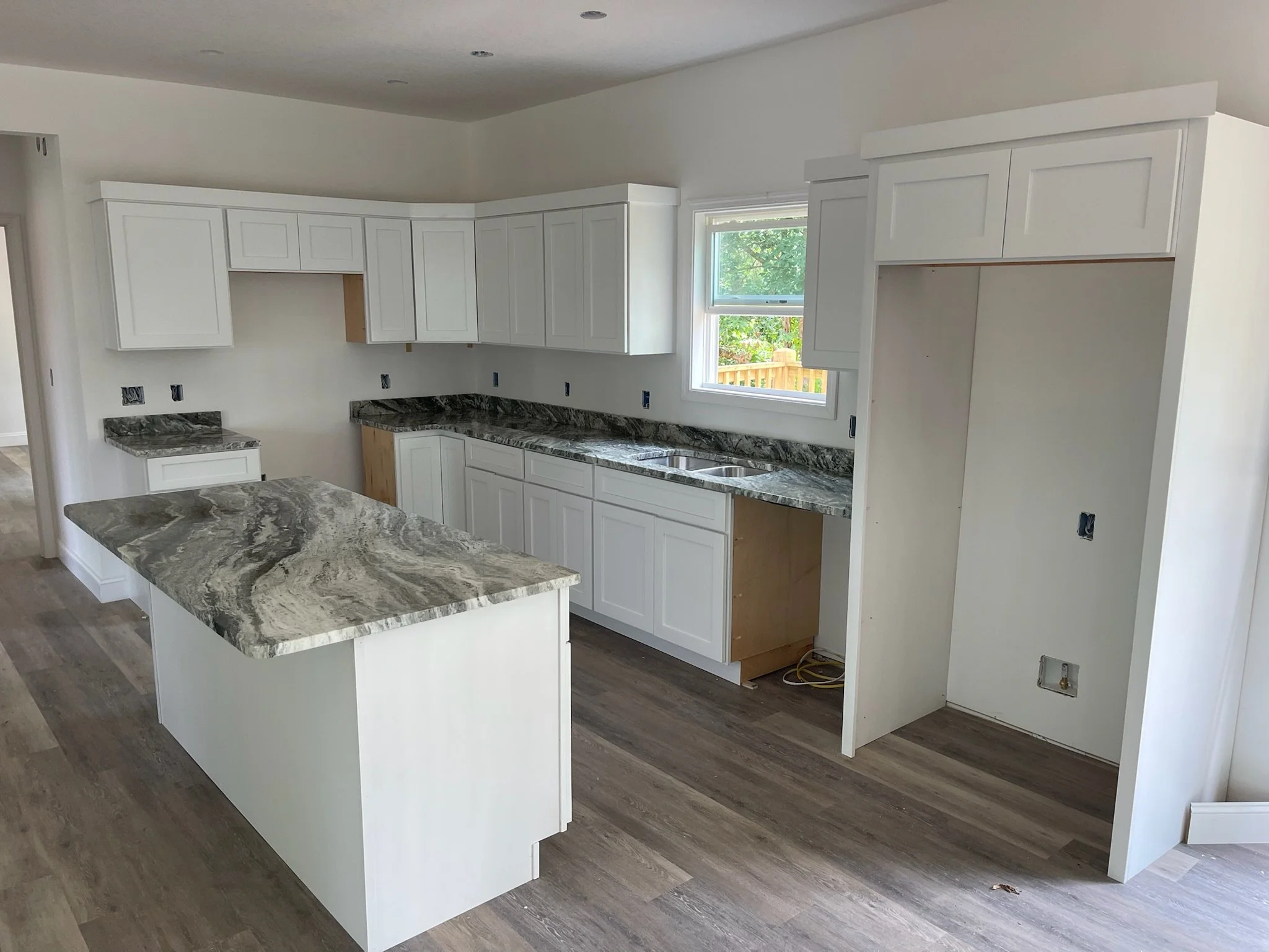 Kitchen under construction with white cabinets, marble countertops, a small island, and an empty space for appliances, with a window overlooking a backyard.