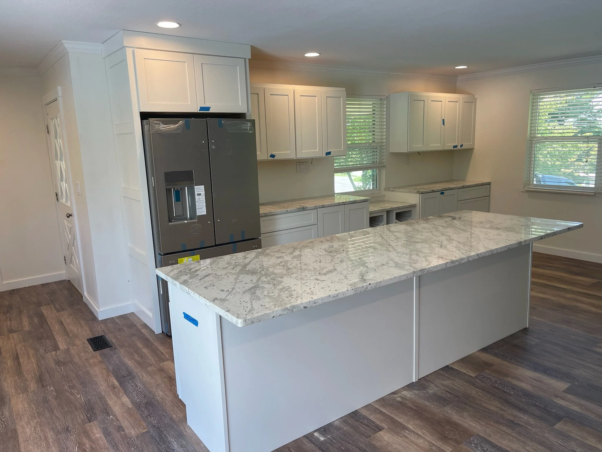 Unfinished kitchen with white cabinets, granite countertops, and a large island, with a stainless steel refrigerator, hardwood flooring, and multiple windows.